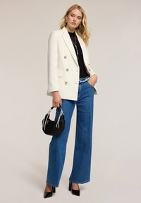 Biała marynarka double-breasted z złotymi guzikami, zestawiona z czarnym golfem i szerokimi niebieskimi dżinsami, trzymająca małą czarną torebkę.