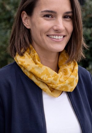 Femme souriante en plein air, portant une écharpe à motifs jaune moutarde, une veste bleu marine et une chemise blanche.