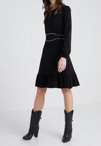 Robe noire à manches longues avec un ourlet à volants, accentuée par une taille ornée de clous. Portée avec des bottes cowboy à talons noires. Texture de tissu lisse.