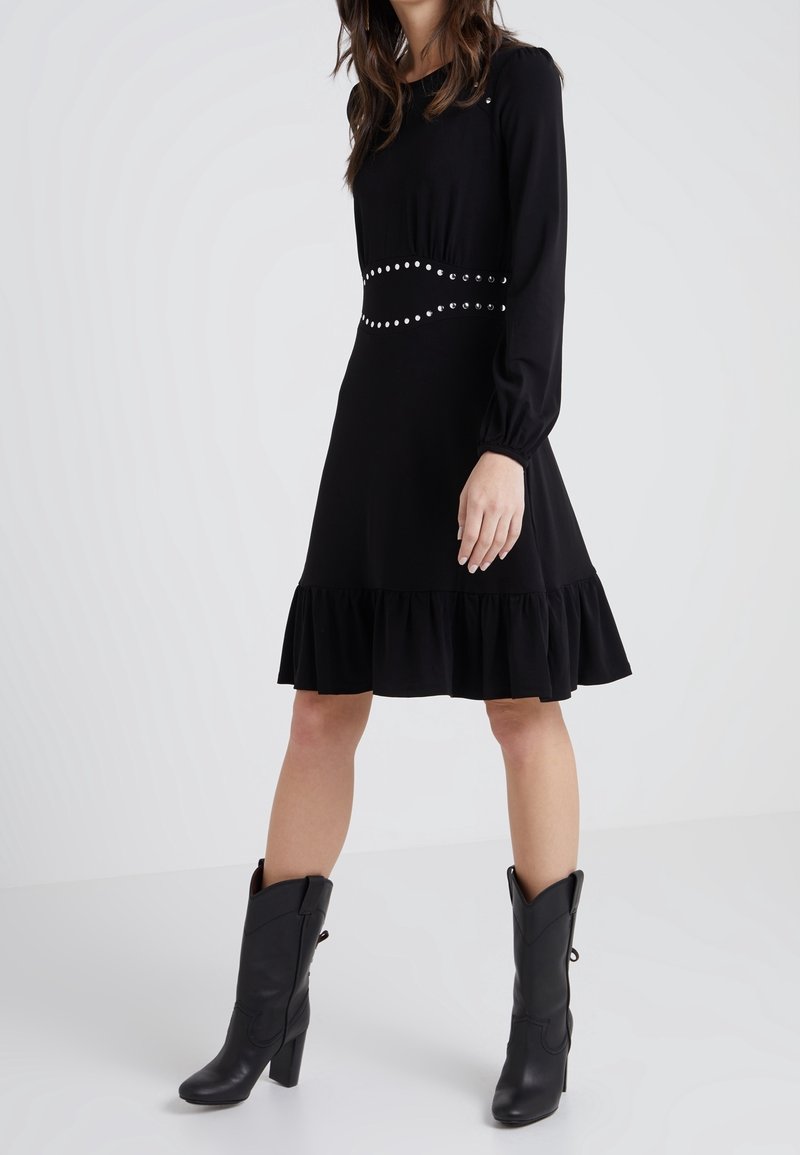 Robe noire à manches longues avec un ourlet à volants, accentuée par une taille ornée de clous. Portée avec des bottes cowboy à talons noires. Texture de tissu lisse.