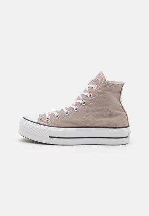 Beige højtliggende lærreds-sneaker med hvid gummisål, hvide snørebånd og sort kant, vist i profil på en ensfarvet hvid baggrund.