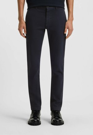 Dark blue cotton trousers with a straight-leg cut; featuring belt loops, front pockets, and a smooth texture. Paired with black lace-up boots.