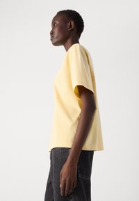T-shirt en coton jaune à manches courtes avec une coupe décontractée, présentant un ourlet droit et un design minimal, associé à un jean foncé.