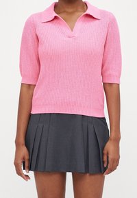 Vaaleanpunainen neulottu polo-paitasweater, jossa on lyhyet hihat, ribbikuvio ja V-pääntie. Yhdistetty tummanharmaisiin laskostettuihin hameeseen.