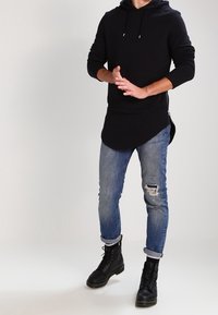 Felpa nera con cappuccio e tasca frontale, indossata sopra jeans blu strappati, abbinata a stivali neri stringati. Tessuto leggero, stile informale.