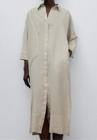 Abito in lino beige con bottoni frontali, colletto e maniche a tre quarti. Presenta un dettaglio laterale testurizzato e asimmetrico e tasche frontali.