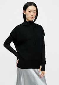 Μαύρος, κοντομάνικος pullover με κουκούλα και κορδόνι, φορεμένος πάνω από ένα μαύρο μπλουζάκι με μακριά μανίκια και μια λαμπερή ασημένια φούστα. Ομοιόμορφη υφή.