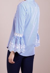 Blå och vit randig skjorta med spetsdetaljerade trumpetärmar, knäppning i kragen och en figursydd design. Mjuk tyg, avslappnad passform.