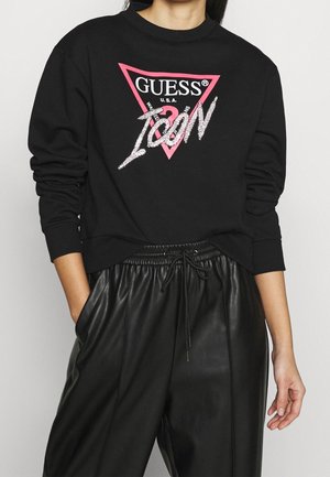 Person trägt schwarzen Pullover mit pinkem und weißem Guess Icon-Logo und schwarze Kunstleder-Jogginghose mit Kordelzug, Hände in den Taschen.