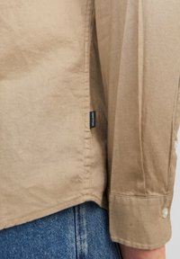 Khaki button-up shirt made of lightweight fabric, featuring a side seam tag and a cuff with white buttons. Blue denim visible below.