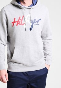 Felpa grigia con tasca frontale, cappuccio con lacci e logo stampato "Hilfiger Denim" in rosso, bianco e blu. Materiale jersey morbido.