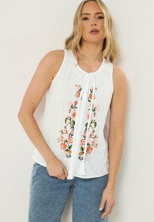 Femme blonde portant un chemisier sans manches blanc avec une broderie florale orange et rose, associée à un jean bleu, debout devant un mur clair.