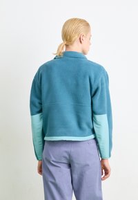 Fleece jacket featuring a teal body with light blue sleeve accents and a collar. The texture is soft and plush with a cropped design.