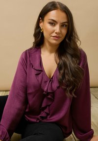 Langärmlige lila Bluse mit V-Ausschnitt und Rüschendetail entlang des Halsausschnitts, kombiniert mit schwarzen Hosen. Glatte Textur und subtiler Glanz.