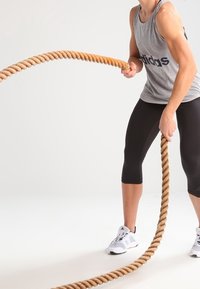 Entrenando con una cuerda de batalla gruesa, retorcida, de color marrón claro, sosteniéndola con ambas manos. La persona lleva una camiseta sin mangas gris y unos leggins negros.