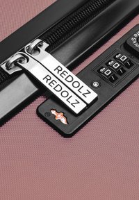Valise rose avec une surface texturée, dotée de doubles tirettes de fermeture éclair en argent, étiquetées "REDOLZ", et d'un cadenas à combinaison sur matériel noir.
