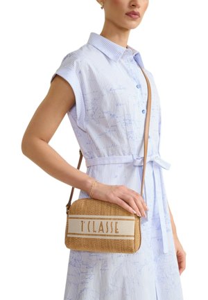 Donna con un vestito azzurro chiaro a maniche corte con cintura, che tiene una borsa a tracolla intrecciata beige con la scritta "T CLASSE" sul davanti.