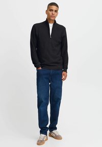 Schwarzer Reißverschluss-Pullover mit einem strukturierten Muster, kombiniert mit blauen geraden Jeans und grauen Sneakern mit weißen Streifen.