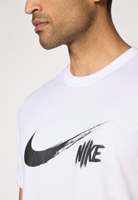 Fehér pamut póló, nagy fekete Nike swoosh szimbólummal és "NIKE" felirattal ecsetvonás stílusban, kerek nyakkivágással és rövid ujjakkal.