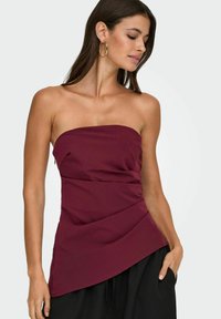 Bordo brezramni top z oprijetim korpusom, z drapirano teksturo, asimetričnim robom in gladkim materialom. Usklajen s črnimi ohlapnimi hlačami.