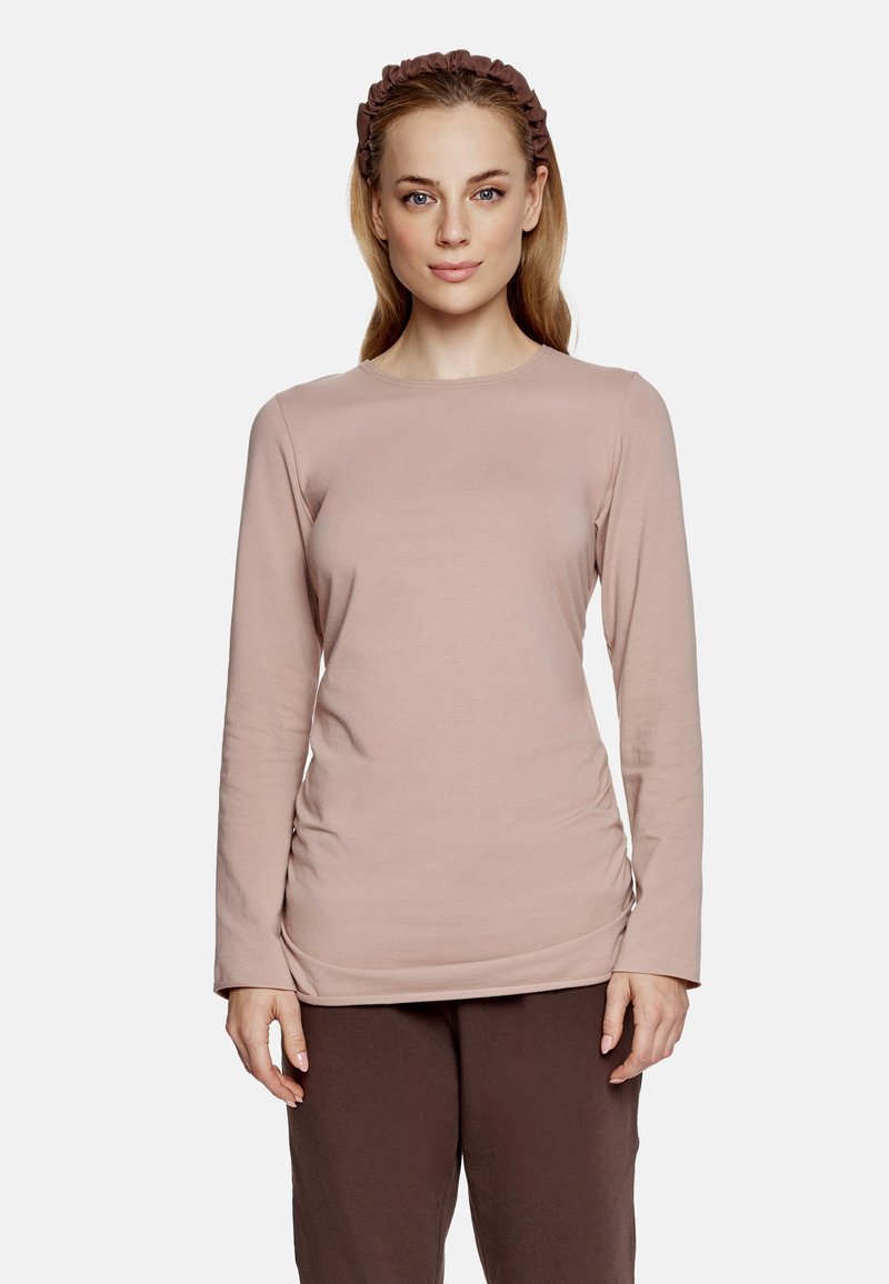 Długi rękaw, top z okrągłym dekoltem w miękkim, jasnoróżowym materiale z marszczonymi detalami po bokach, w połączeniu z brązowymi spodniami i pasującą opaską na głowę.