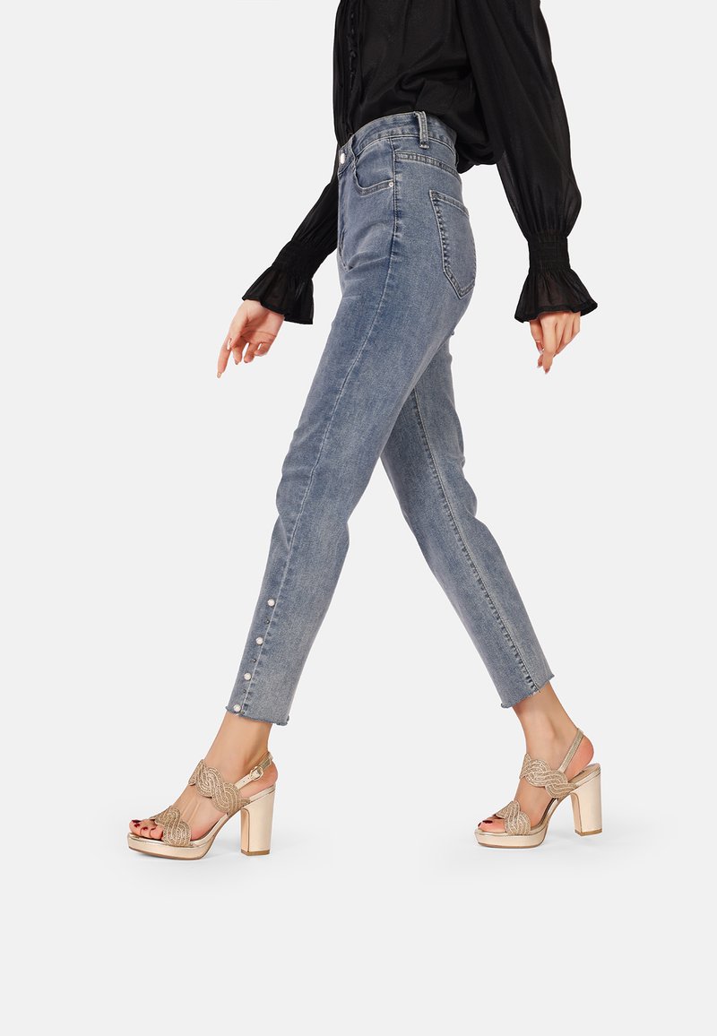 Jeans in denim azzurro chiaro con dettagli a bottone sulla caviglia, abbinati a sandali beige con tacco a pianta testurizzata e un design di fascia decorativa.