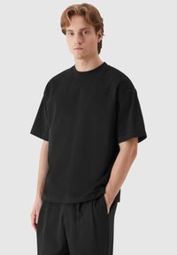 Fekete, oversized póló, rövid ujjú, kerek nyakú, pamutból készült. Simaszerű textúra, laza szabás, laza fekete nadrágokkal párosítva.