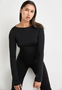 Svart langermet bodysuit med rund hals, tettsittende design og myk tekstur, kombinert med svarte bukser og et sølvkjede som tilbehør.
