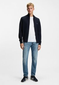 Chaqueta azul marino con cremallera y cuello alto, combinada con una camiseta blanca, jeans azules y zapatillas negras con detalles blancos. Fondo neutro.