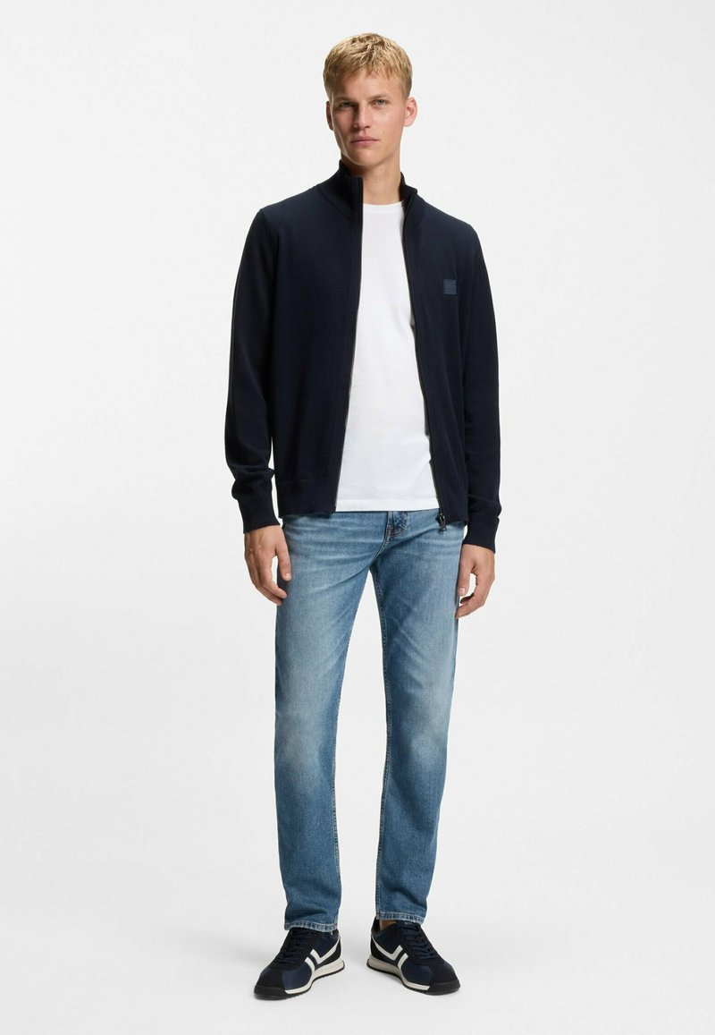 Chaqueta azul marino con cremallera y cuello alto, combinada con una camiseta blanca, jeans azules y zapatillas negras con detalles blancos. Fondo neutro.