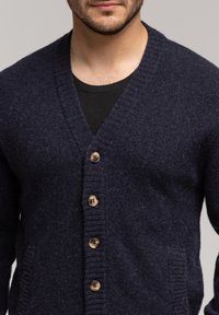 Granatowy kardigan wełniany z dekoltem w kształcie litery V, pięcioma brązowymi zapięciami na guziki, ribbowymi mankietami i dwiema bocznymi kieszeniami. Teksturowana tkanina z delikatnym meszkiem.