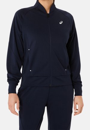 Person wearing a navy blue zip-up jacket and matching pants with Asics logo on the left chest, standing with arms relaxed by their sides.