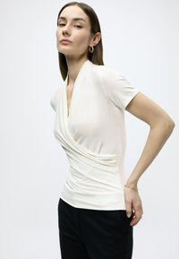 Λευκό τοπ με κοντά μανίκια, με draped, τύπου φάκελου εμπρός και λεία υφή, σε συνδυασμό με μαύρο παντελόνι. Μινιμαλιστικός σχεδιασμός, χωρίς ορατά σχέδια.