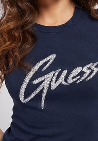 Námořnická modrá mikina vyrobená z měkkého materiálu, s texturovaným, třpytivým stříbrným logem "Guess" na hrudi a klasickým kulatým výstřihem.