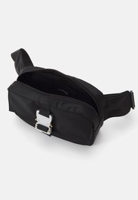Juodas audinys fanny pack'as su užtraukiama kišenė, sidabriniu užsegimu ir reguliuojama dirželiu; lygus paviršius ir minimalizmo stilius.