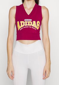 Bordó bezramenný crop top s výstřihem do V, zdobený velkým žlutým a bílým logem a textem "ADIDAS", v kombinaci s bílými legínami s vysokým pasem.