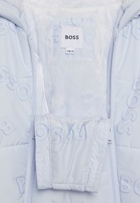 Světlá modrá prošívaná bunda s chlupatou vnitřní podšívkou, zdobená vzorem s logem BOSS a zapínáním na zip. Štítek označený "BOSS" s informacemi o velikosti.