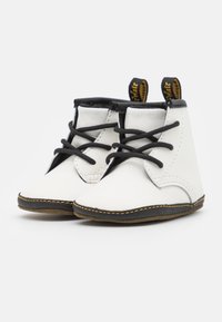 Botitas de bebé blancas con cordones negros, ribetes negros, costuras amarillas y lengüetas con letras amarillas sobre un fondo blanco.