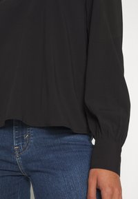 Blusa nera con vestibilità ampia e maniche a sbuffo, abbinata a jeans blu. Il tessuto sembra liscio con una leggera lucentezza e una sottile caduta.