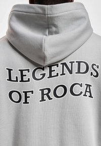 Grå hætte-trøje med teksten "LEGENDS OF ROCA" i fed sort skrift på ryggen, lavet af blødt, struktureret stof.