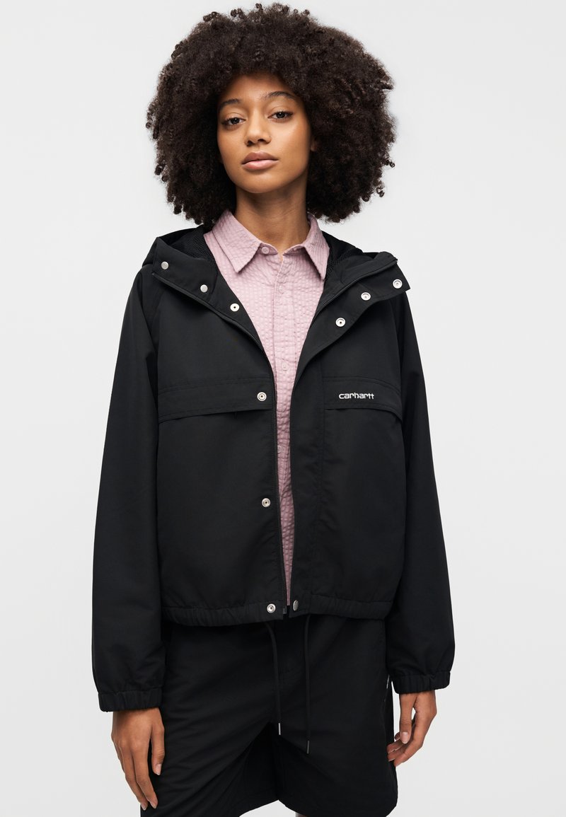 Giacca a vento nera con cappuccio, bottoni automatici sul davanti, due tasche sul petto e polsini elasticizzati, indossata sopra una camicia rosa con collo.