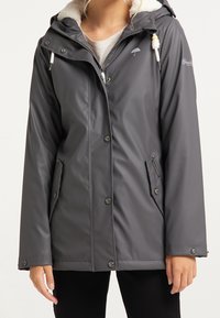 Woman wearing a dark gray waterproof jacket with snap buttons, front zipper, pockets, and a hood with white drawstrings and fleece lining.
