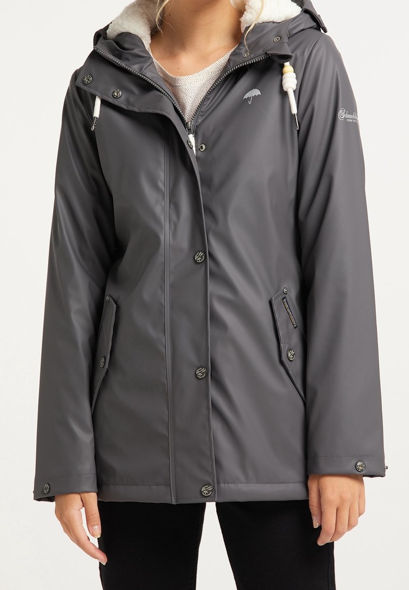 Woman wearing a dark gray waterproof jacket with snap buttons, front zipper, pockets, and a hood with white drawstrings and fleece lining.