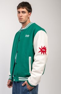 Zelený a biely varsity kabát s výšivkou. Má zelené telo, biele kožené rukávy, rebrované manžety a červený vyšitý vzor na rukáve.