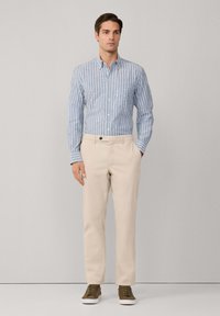 Lichtblauw en wit gestreept overhemd met knopen, beige broek en olijfgroene sneakers. De broek heeft een zak en een knopenaccent.