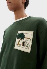 Felpa verde con una toppa testurizzata raffigurante un ricamo di un albero e un edificio, etichettata "Modalità Mediterranea."