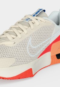 Μπεζ αθλητικό παπούτσι Nike με υφή στο επάνω μέρος, λευκό λογότυπο swoosh, κρεμ κορδόνια και κόκκινο-πορτοκαλί λεπτομέρειες στη σόλα.