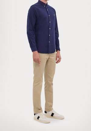 Mand iført navyblå skjorte med knapper, beige bukser og hvide sneakers med sorte striber, stående mod en ensfarvet hvid baggrund.