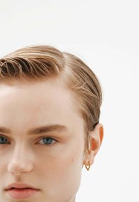 Pendiente de aro dorado con un diseño en forma de corazón, lucido en una oreja perforada. El modelo tiene el cabello corto y peinado, y un fondo neutral.
