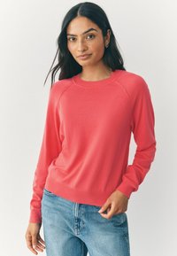 Korallfarbener Strickpullover mit rundem Halsausschnitt, gerippten Bündchen und Saum. Weiche Textur und Raglanärmel, kombiniert mit blauen Jeans.